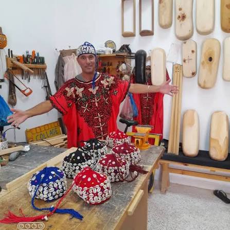 Maalem Hassan in vibrant traditional red embroidered dress with arms open in front of carved guembri soundboxes in his Essaouira workshop